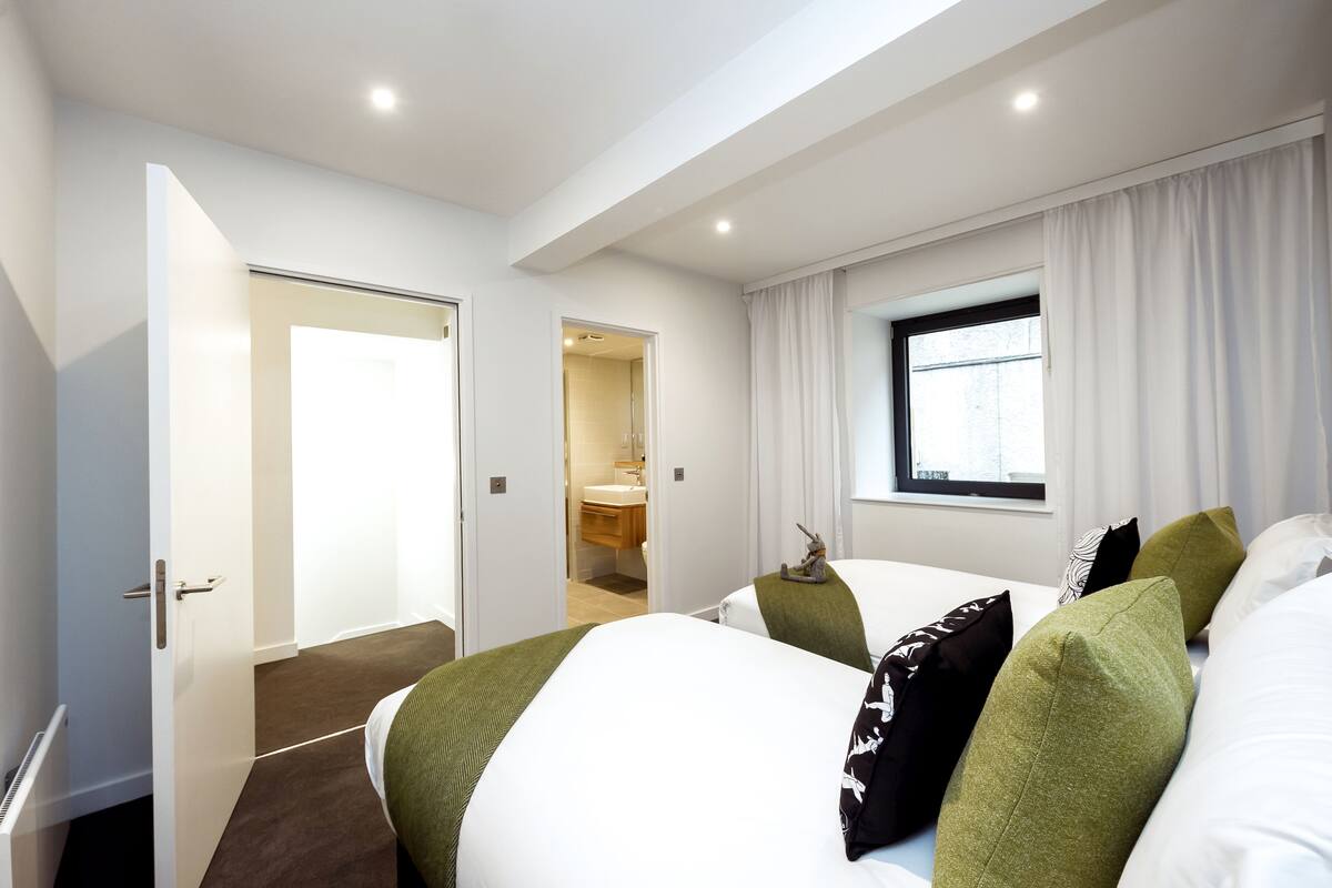 A hotel room with two beds, a window with curtains, a bathroom visible through an open door, and a ceiling light.