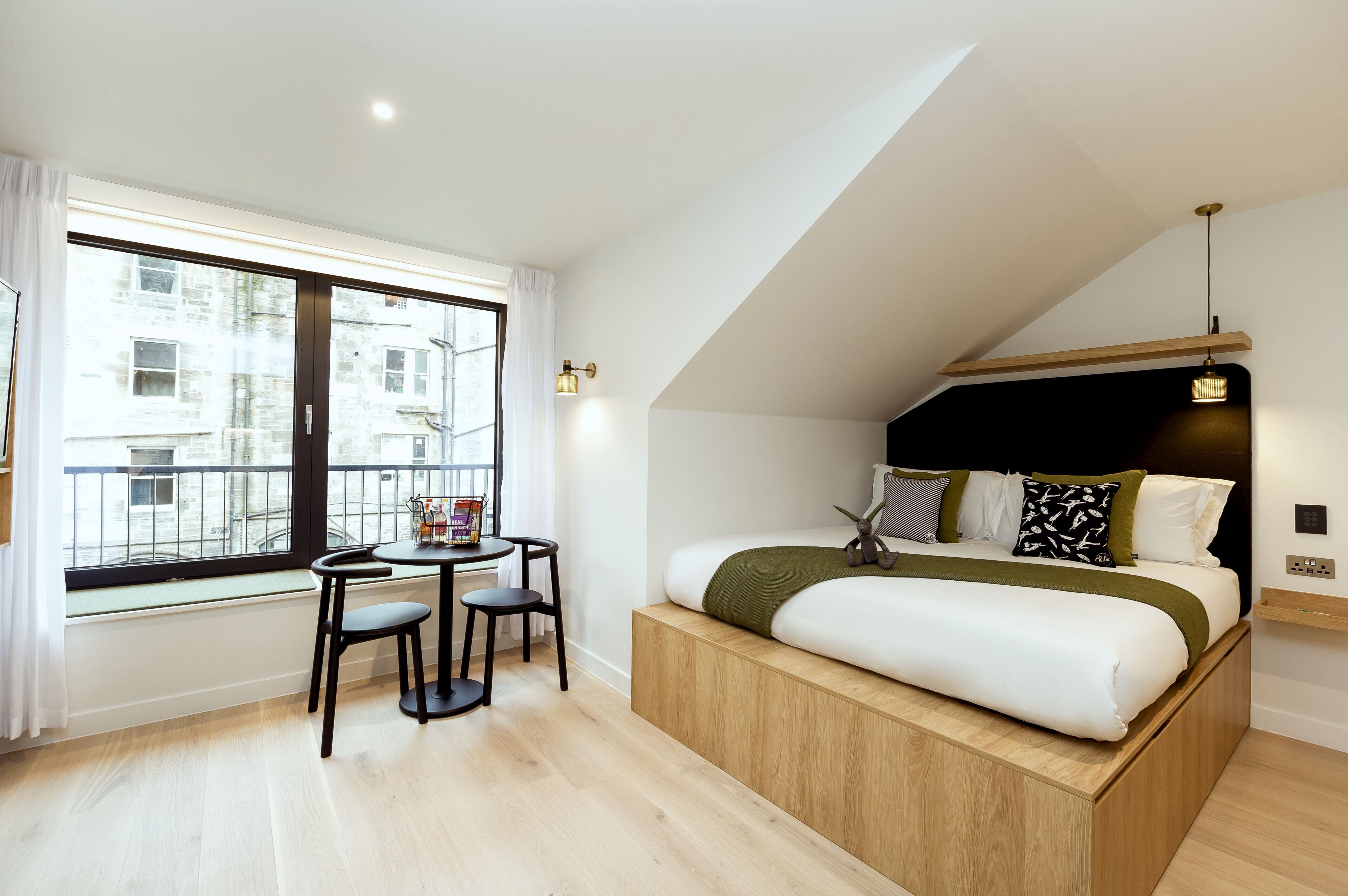 Un dormitorio moderno con cama alta, una mesita con sillas y un amplio ventanal que ofrece vistas a la ciudad.