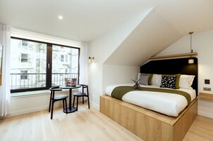 Un dormitorio moderno con cama alta, una mesita con sillas y un amplio ventanal que ofrece vistas a la ciudad.