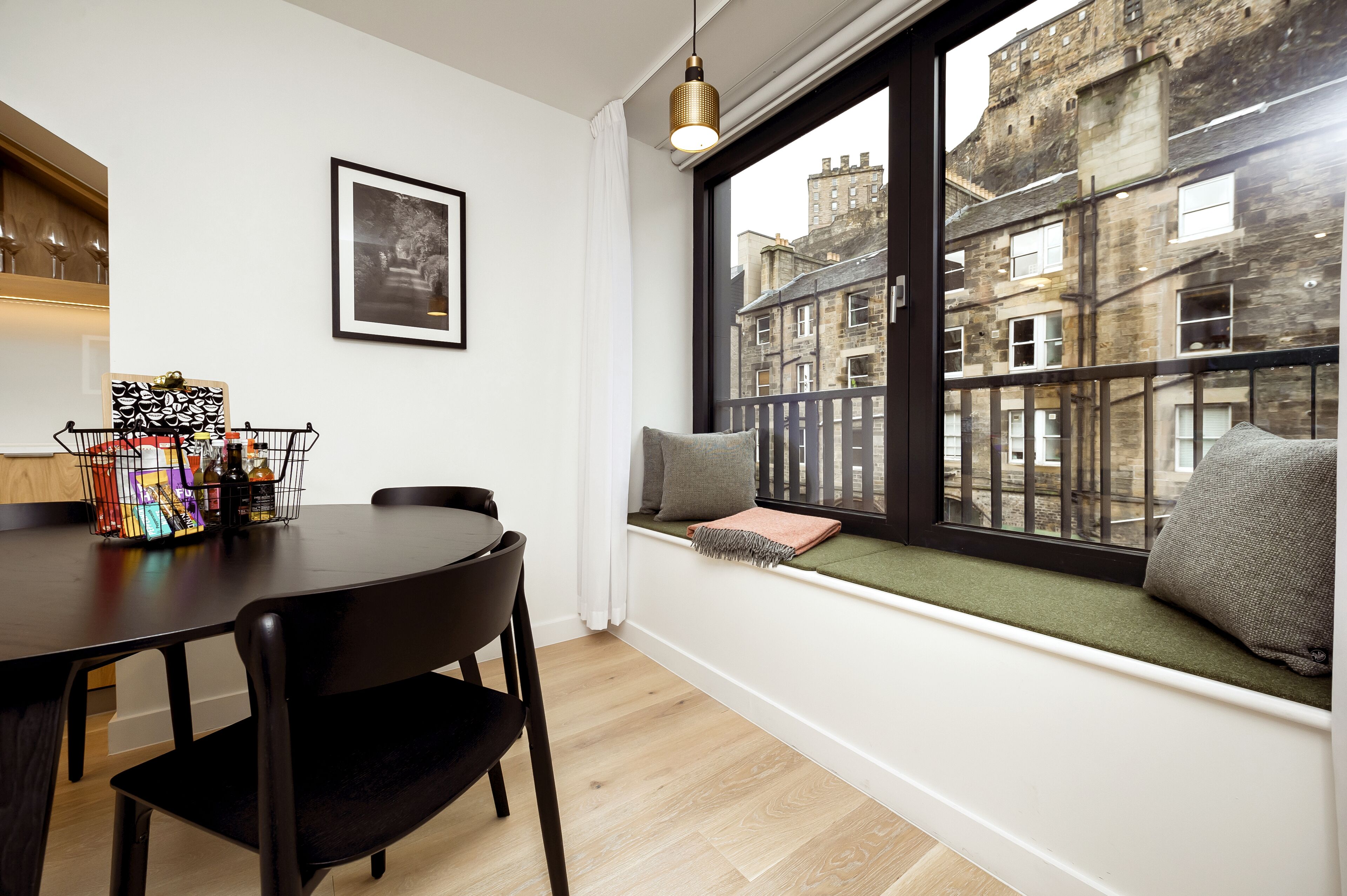 A modern room with a dining table, chairs, a window seat with cushions, a framed picture, and a hanging light fixture.