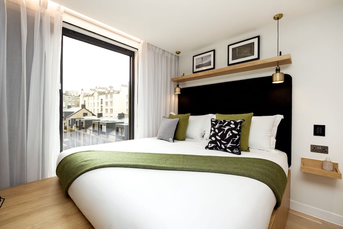 Ein modernes Hotelzimmer mit einem großen Bett, Blick auf Gebäude durch ein Fenster und einer Holzeinrichtung mit einer Lampe.