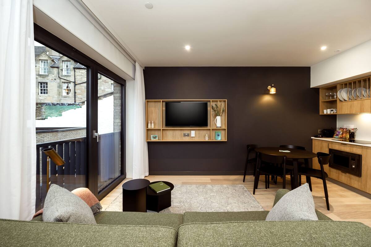 A modern living room with a large window, a flat-screen TV mounted on the wall, a dining area with a table and chairs, and a kitchen area with wooden cabinets and a countertop.