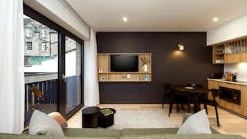Un salon moderne doté d’une grande fenêtre, d’un téléviseur à écran plat fixé au mur, d’un coin repas avec une table et des chaises, et d’un espace cuisine avec des meubles en bois et un plan de travail.