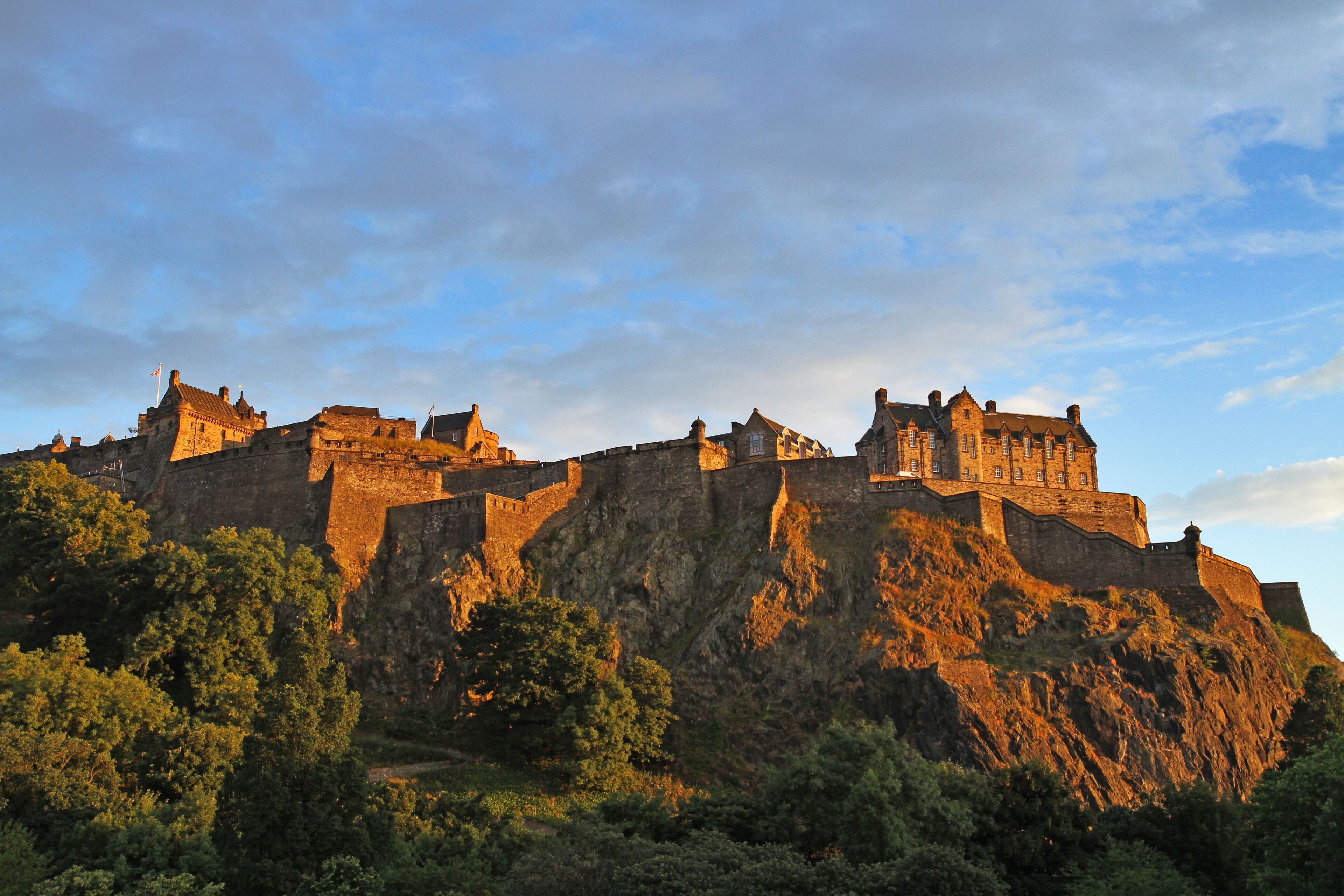 A historic castle with multiple towers and walls, situated on a rocky hill with dense greenery in the foreground.