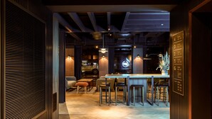 Zona de bar moderna con barra de madera y taburetes, un área de estar con sofá y reposapiés, y un letrero de neón sobre la barra.