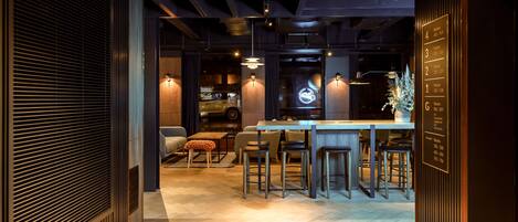 Zona de bar moderna con barra de madera y taburetes, un área de estar con sofá y reposapiés, y un letrero de neón sobre la barra.