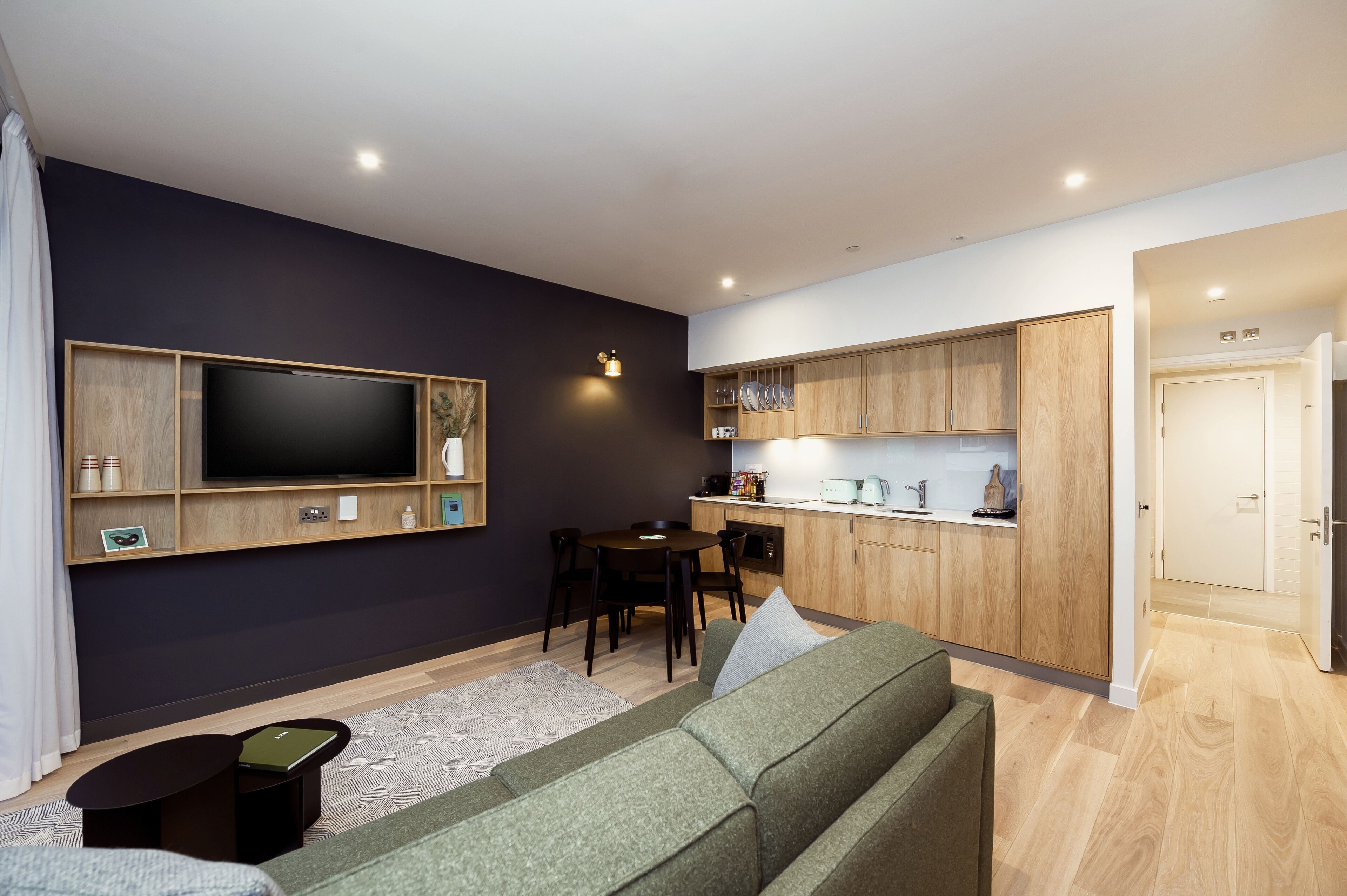 Ein modernes Wohnzimmer mit Flachbildfernseher, einem grünen Sofa, einem kleinen Couchtisch und einem Essbereich mit Tisch und Stühlen.
