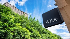Un cartel con el nombre 'Wilde' sobre un fondo de vegetación exuberante y un edificio histórico bajo un cielo azul.