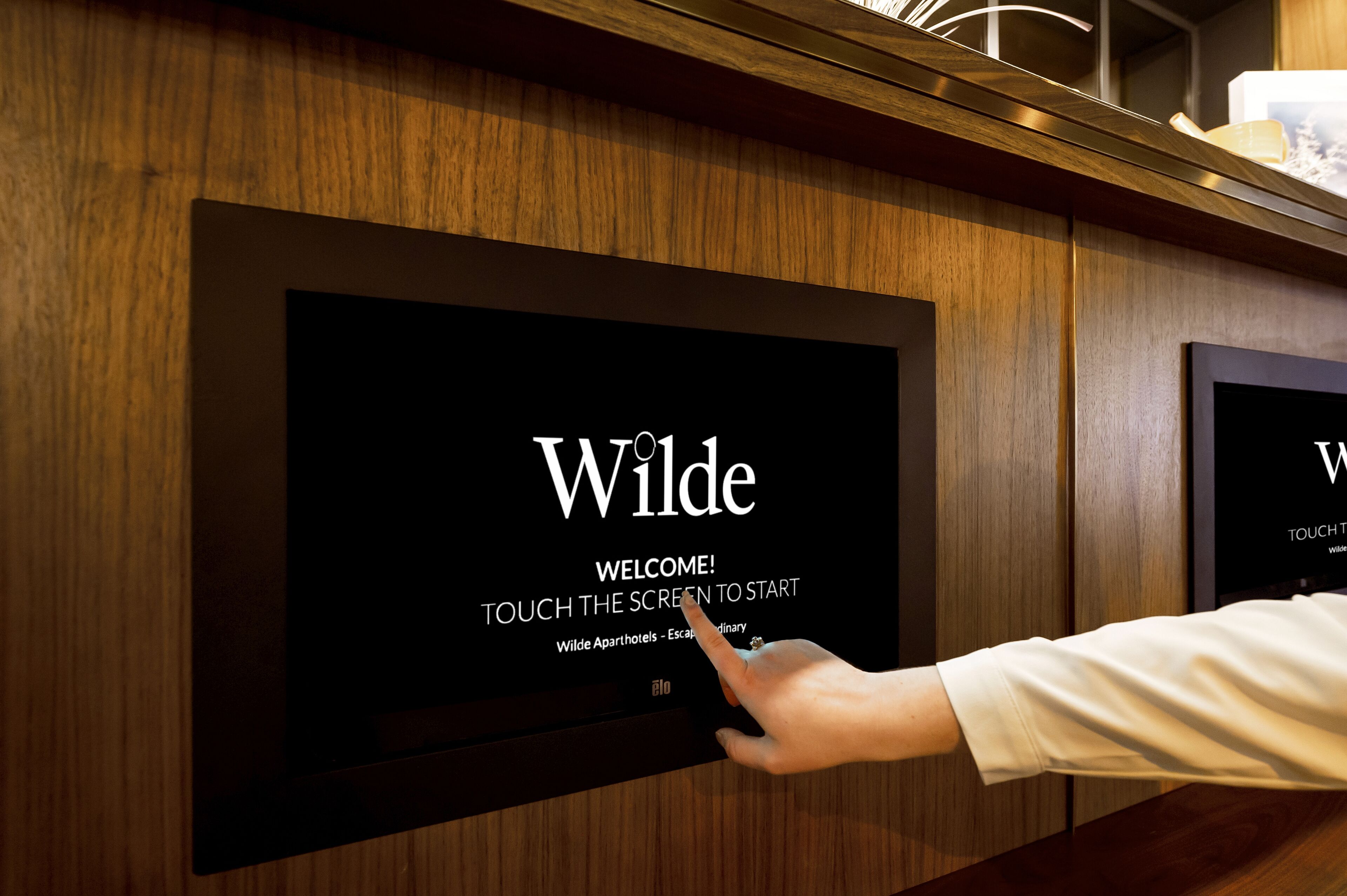 Una mano toca una pantalla que muestra 'Wilde' con un mensaje de bienvenida e instrucciones para iniciar tocando la pantalla.