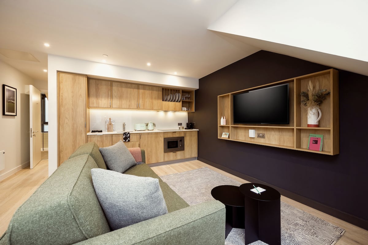 A modern living room with a green sofa, a flat-screen TV mounted on the wall, and a kitchen area with wooden cabinets and a microwave.
