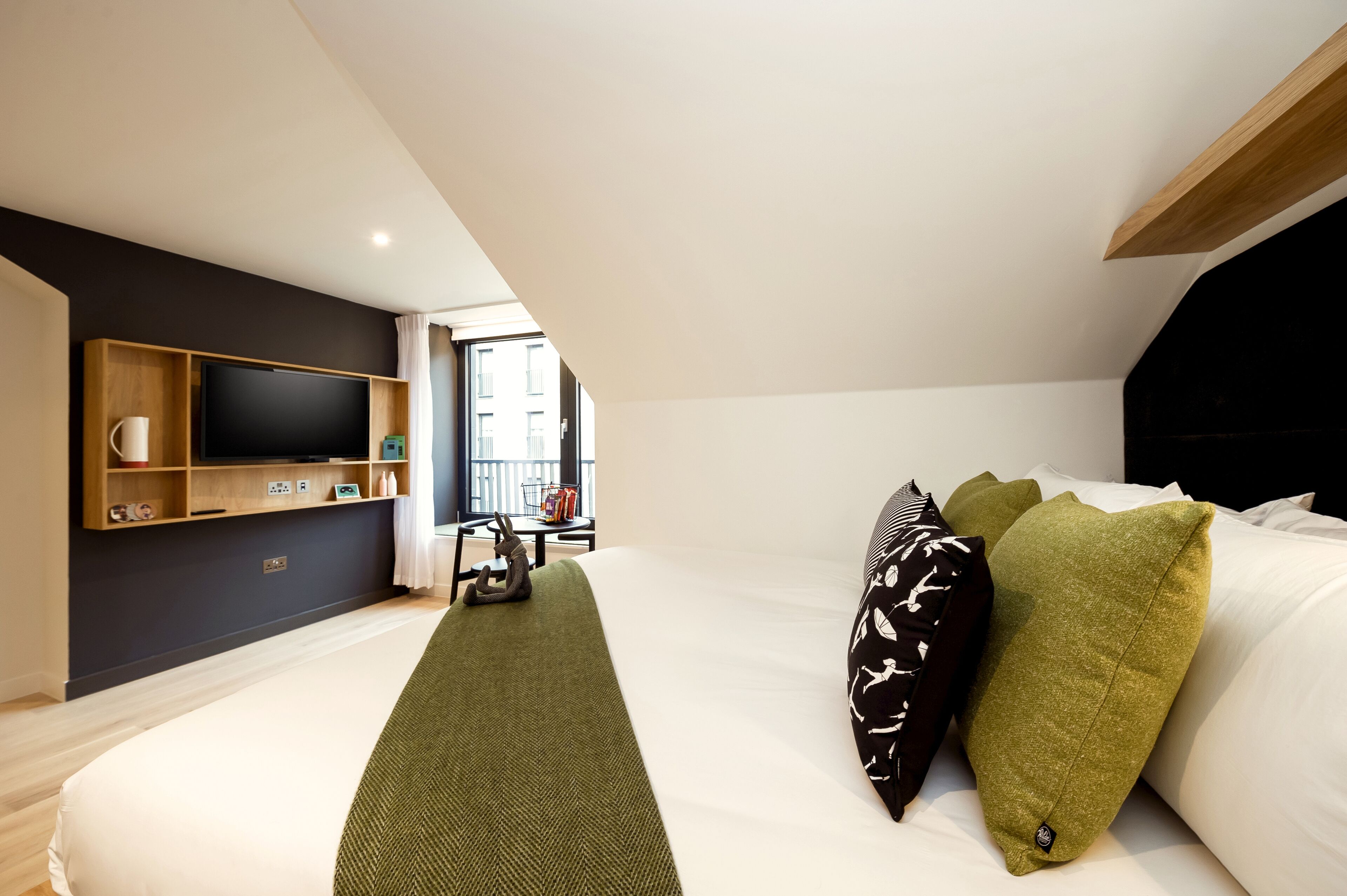 A modern hotel room with a large bed, a flat-screen TV mounted on the wall, a small table with a chair, and a window providing a view of the outside.