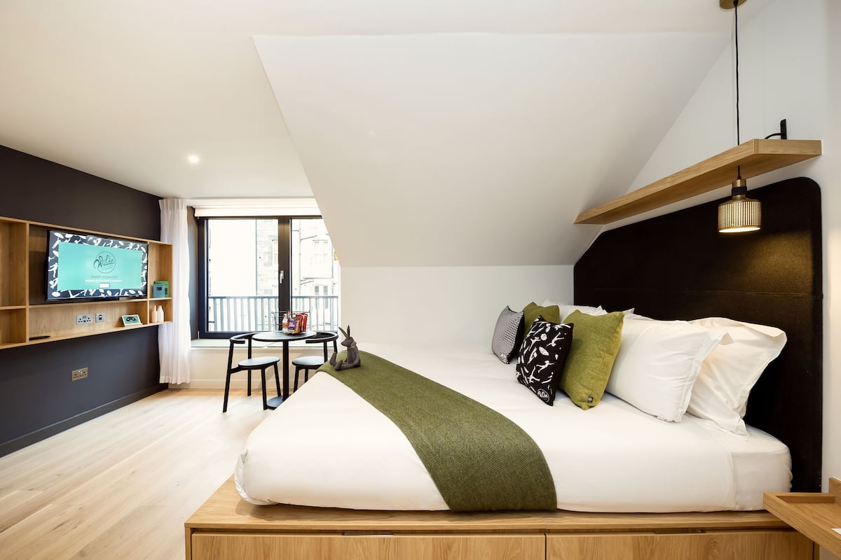 A modern bedroom with a large bed, a TV on a wooden shelf, a small table, and a balcony view.