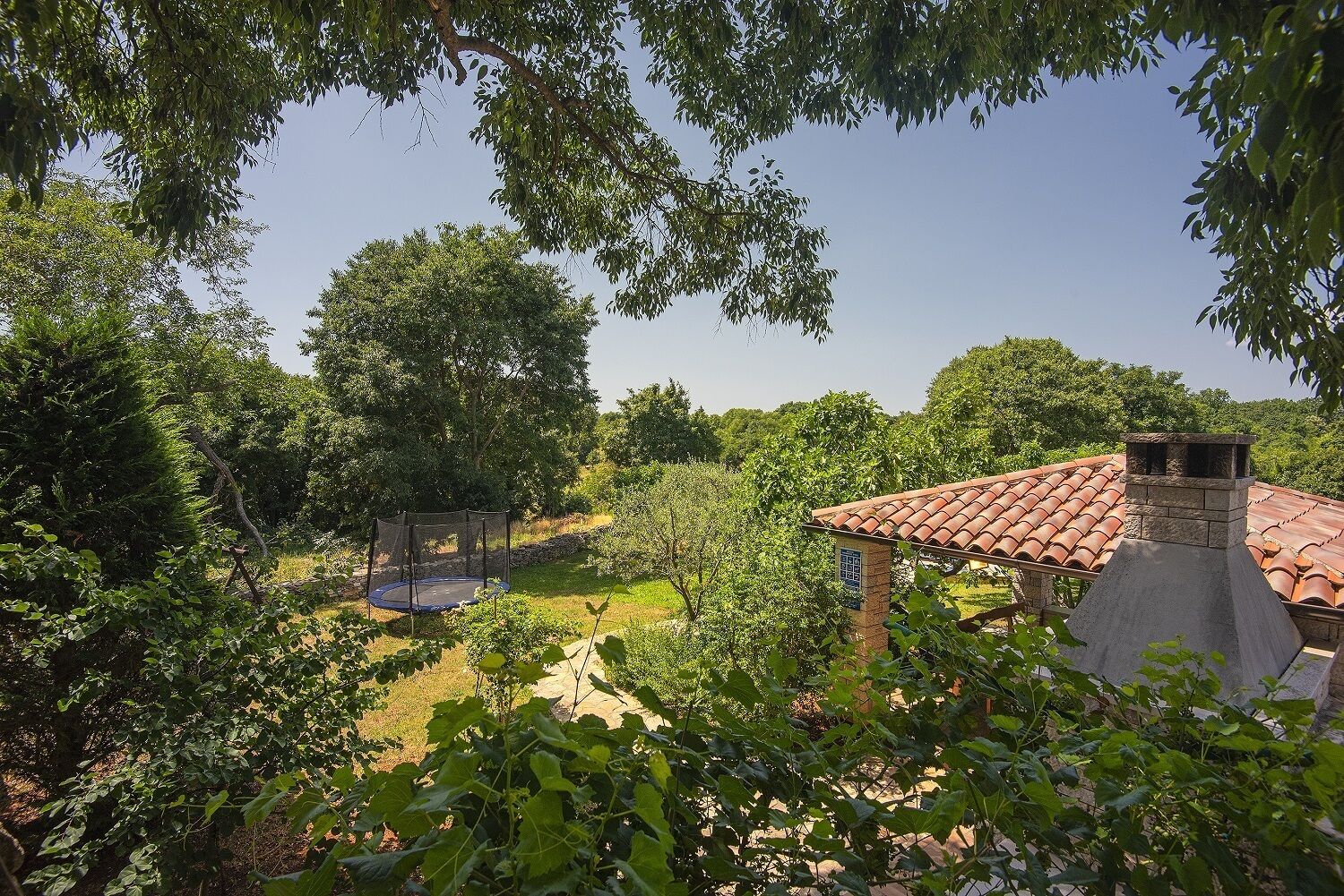 A residential area with a house featuring a tiled roof and a chimney, surrounded by lush greenery and a trampoline in the yard.