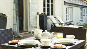 A breakfast setup with plates, cups, and a teapot on an outdoor table, with a house and a garden chair in the background. - Luxury 5-star single-storey cottage for two near the Cornish coast (Helston)
