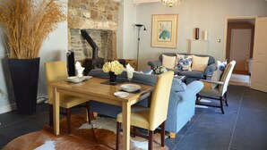 A cozy living room with a stone fireplace, a dining table with chairs, and a sofa with cushions. - Luxury 5-star single-storey cottage for two near the Cornish coast (Helston)
