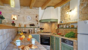 Une cuisine aux murs en pierre, avec des poutres apparentes, un dosseret carrelé et un coin repas où l’on sert des croissants, du jus d’orange et une cafetière.