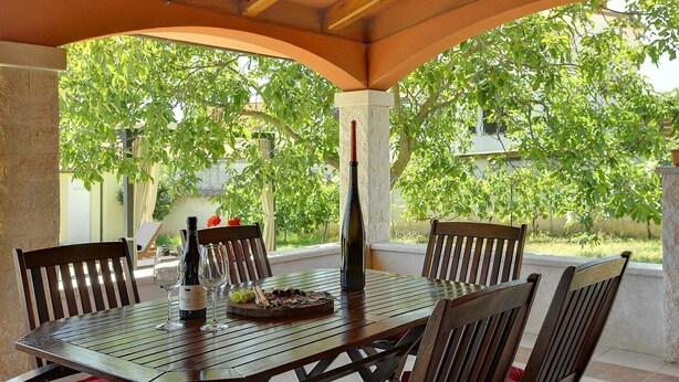 A covered outdoor dining area with a table set for two, surrounded by chairs, and a bottle of wine on the table. - Sunny villa 500 meters to beach with private pool (Peroj)