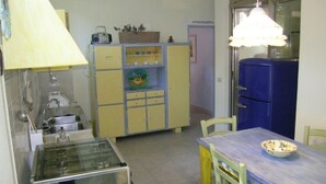 Une cuisine de taille réduite, équipée d’une cuisinière à gaz, de meubles de rangement jaunes, d’un réfrigérateur bleu et d’une table à manger avec des chaises.