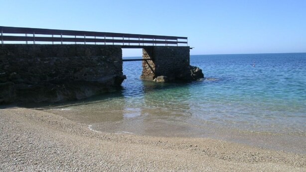 Un pont de pierre enjambe une mer calme et mène à une plage de galets.