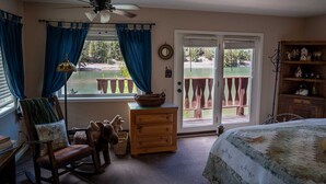 A bedroom with a bed, a dresser, a chair, a desk, a lamp, a ceiling fan, and a view of a deck and lake.