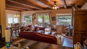 A cozy living room with a red leather sofa, wooden dining table, and chairs, a fireplace, and large windows with a view of the outdoors.