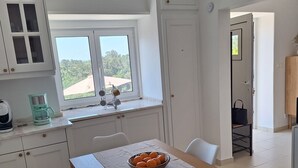 A modern kitchen with white cabinets, a dining table with chairs, a bowl of fruit, and a view of greenery outside.