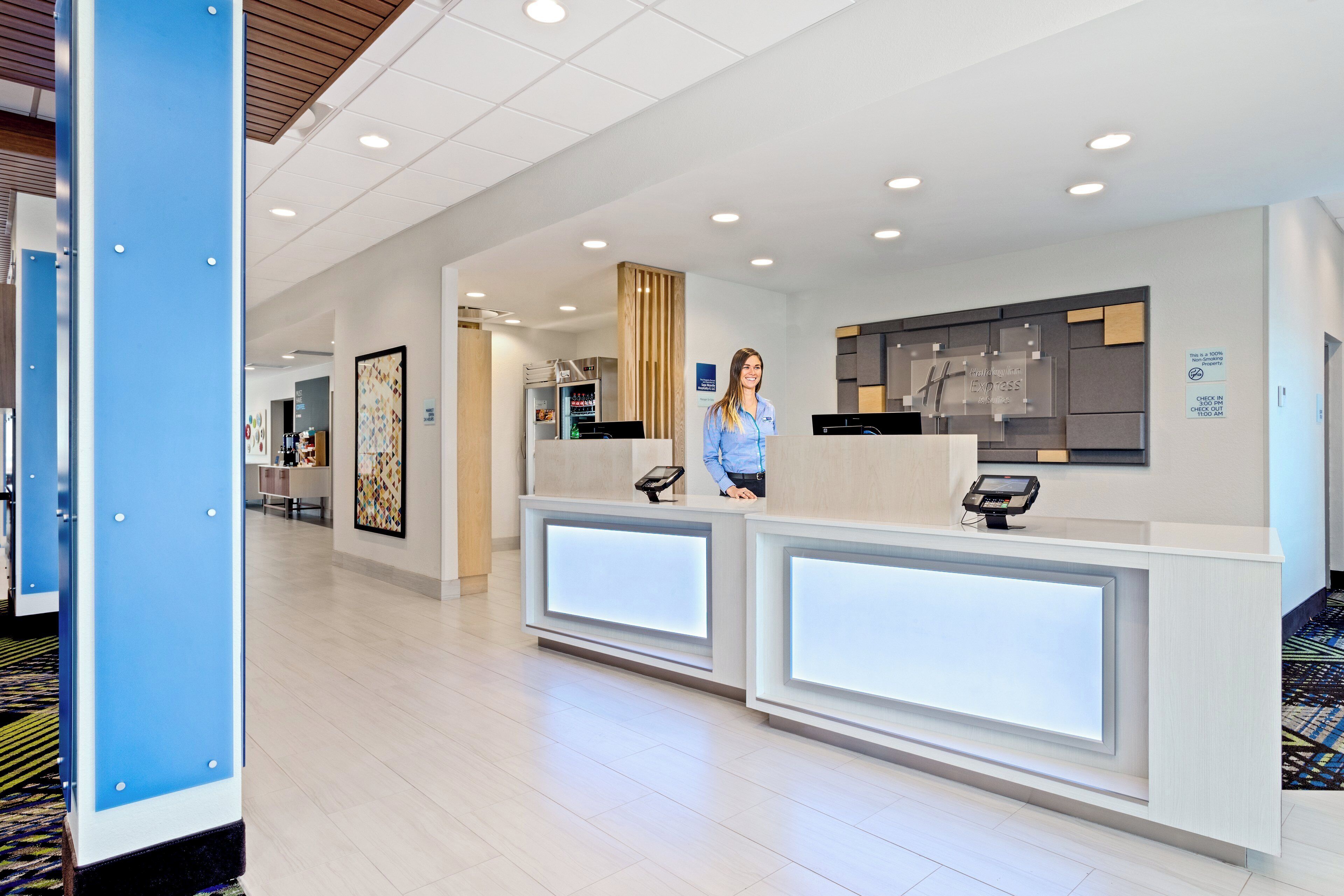 A hotel reception area with a modern design, featuring a reception desk, a digital check-in kiosk, and a wall-mounted sign indicating check-in and check-out times.