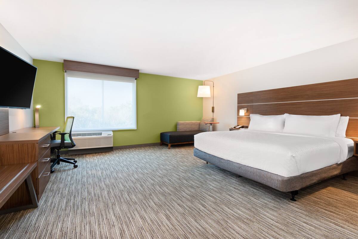 A hotel room with a large bed, a desk with a chair, a TV, a window with blinds, and a lamp.