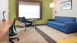 A hotel room with a wooden desk, a blue sofa, a patterned rug, a small table, and a window with blinds. - Holiday Inn Express & Suites Niceville - Eglin Area by IHG (Niceville)