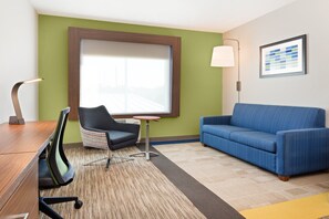 A hotel room with a wooden desk, a blue sofa, a patterned rug, a small table, and a window with blinds. - Holiday Inn Express & Suites Niceville - Eglin Area by IHG (Niceville)