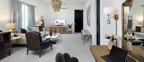 A modern living room with a sofa, armchairs, a coffee table, and a TV. There is a laptop on a wooden table, and a mirror on the wall.