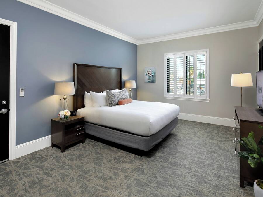 A hotel room with a bed, nightstands, a desk, a television, and a window with blinds.