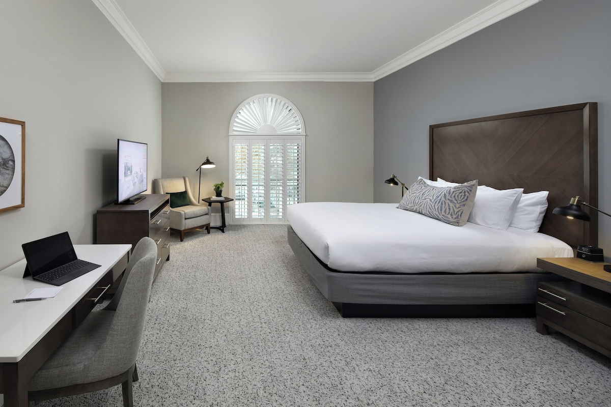 A hotel room with a bed, desk, chair, and a large window with blinds.