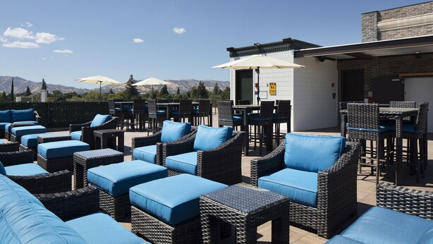 An outdoor seating area with wicker furniture and blue cushions, a bar area with umbrellas, and a view of mountains in the background. - Hotel Winters (Winters)