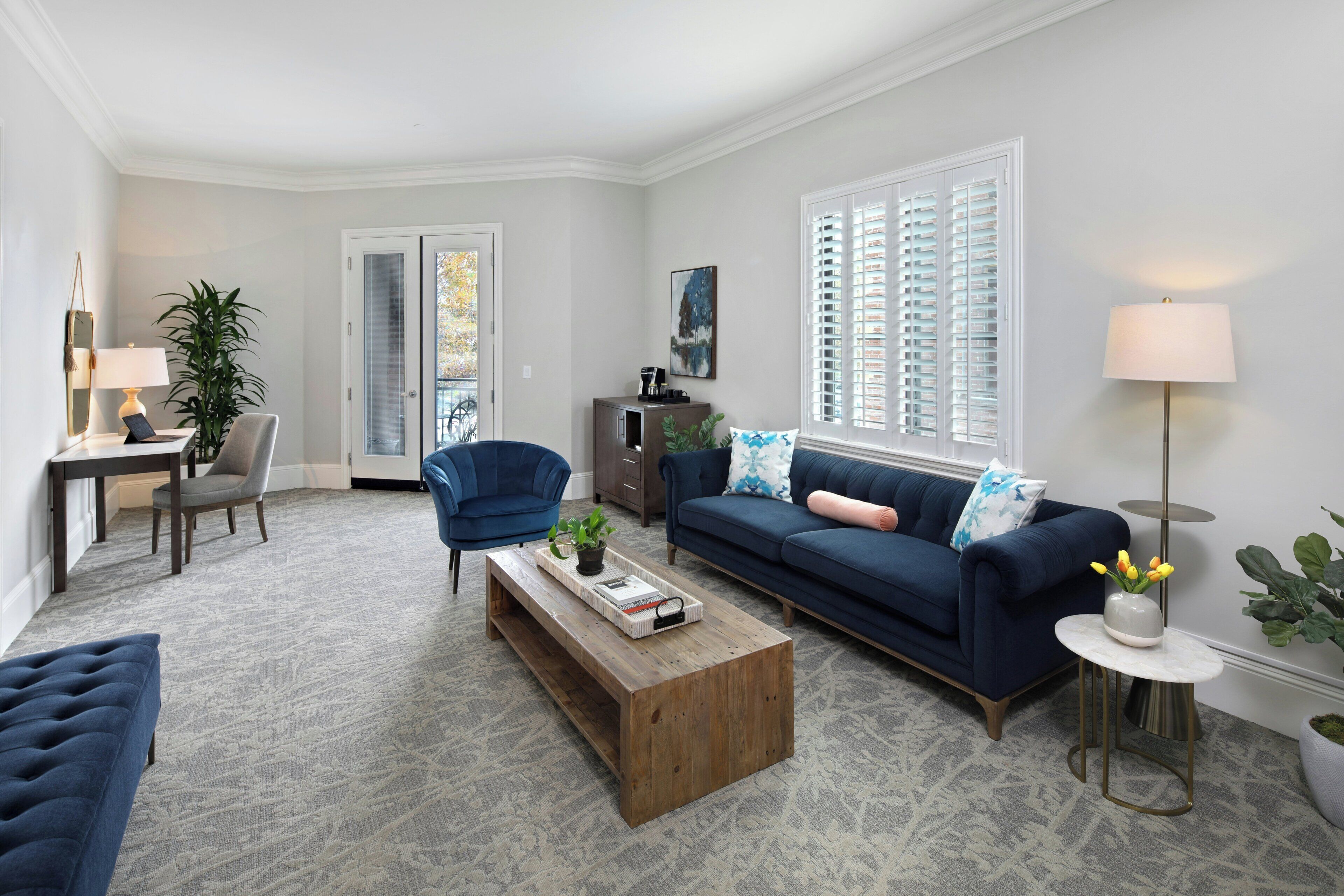 A living room with a blue sofa, a wooden coffee table, a blue armchair, a desk with a lamp, and a large window with blinds.