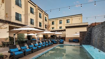 An outdoor pool area with lounge chairs and umbrellas, a barbecue setup, and a stone wall.
