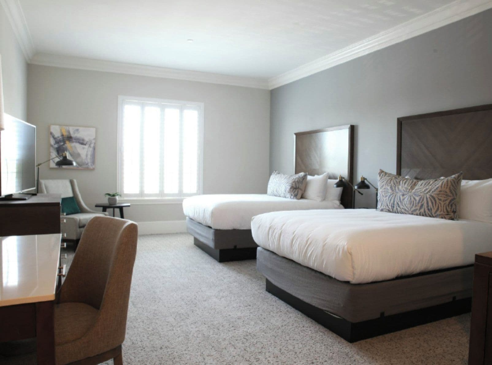 A hotel room with two beds, a desk, a chair, a television, and a window.