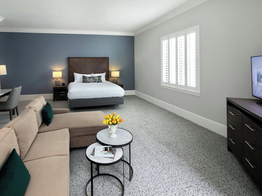 Ein Hotelzimmer mit Bett, Sofa, kleinem Tisch mit Blumenvase, Schreibtisch mit Lampe und Fernseher auf einem Schrank.