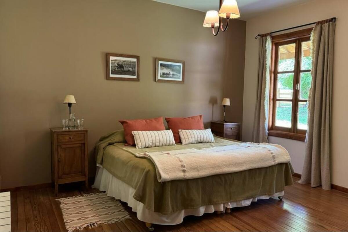 Quarto com cama, duas mesas de cabeceira, janela com cortinas e duas luminárias na parede.