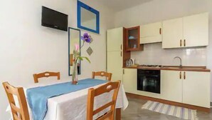 A dining area with a table and chairs, a kitchen with cabinets and a stove, and a television mounted on the wall. - Signorino Resort Quadruple Studio Apartment (MARSALA)