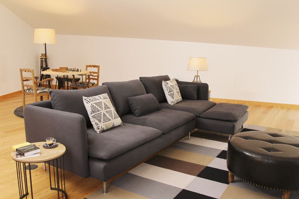 Un salon moderne avec un canapé d’angle, une table basse ronde et un pouf noir. On aperçoit à l’arrière-plan un coin repas avec une table et des chaises.