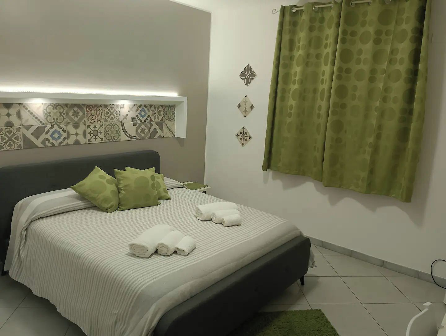 Ein Schlafzimmer mit einem Bett, Kissen, einem Nachttisch, einer Wandleuchte, dekorativer Wandgestaltung mit Muster und einem grünen Vorhang.