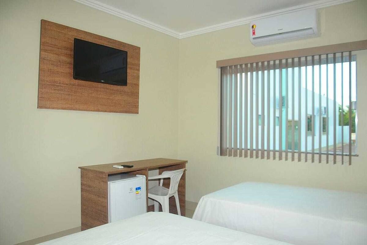 Habitación de hotel con dos camas, un escritorio con computadora, un televisor montado en la pared y una ventana con persianas.
