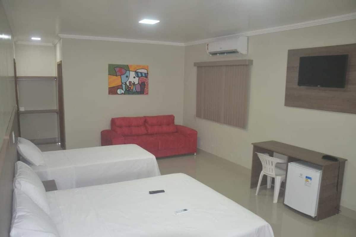Habitación de hotel con dos camas, un sofá rojo, un escritorio con silla, un refrigerador pequeño y un televisor montado en la pared.