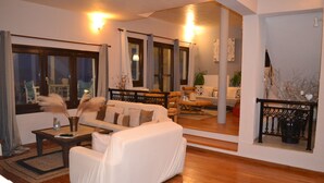 A living room with a sofa, a coffee table, and a dining table. There is a large window with curtains, a plant, and a mirror reflecting the room. - "Villa Erifili" Luxurious villa with spectacular view in Skiathos Island (Taxiarchis)