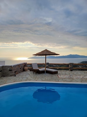 Un espace piscine équipé de chaises longues et d’un parasol, offrant une vue panoramique sur la mer et les montagnes.