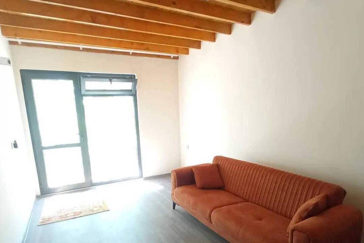A spacious living room with a wooden ceiling, a large sliding glass door leading to a bright area, and a single orange sofa.