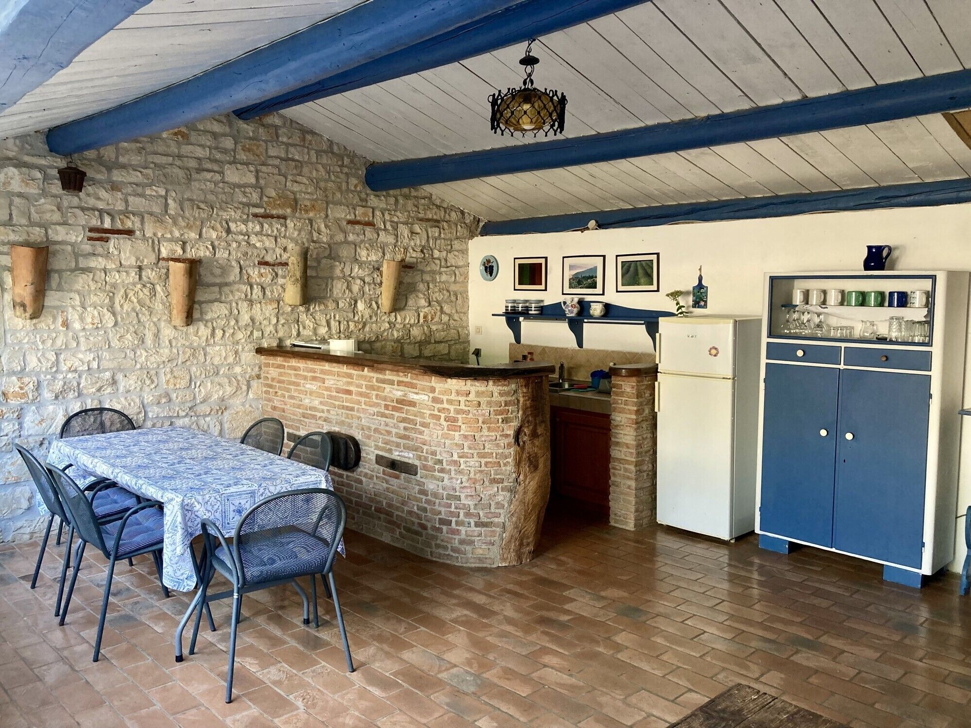 Una cocina rústica con pared de piedra, una mesa de madera con sillas, un armario azul y una lámpara colgante.