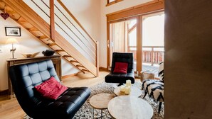 Uma sala de estar ampla com um sofá de couro preto, um travesseiro vermelho e uma mesa de centro redonda. Uma escada de madeira leva a uma varanda com corrimão de madeira.