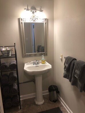 Ein Badezimmer mit einem Standwaschbecken, einem Spiegel mit Zierleuchten, einem Handtuchhalter mit Handtüchern und einem Mülleimer.